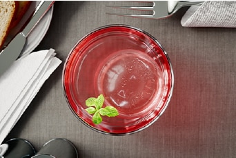 A glass with LG Craft Ice® and a sprig of mint placed on a darken wooden table, alongside utensils, napkins, a plate of food.